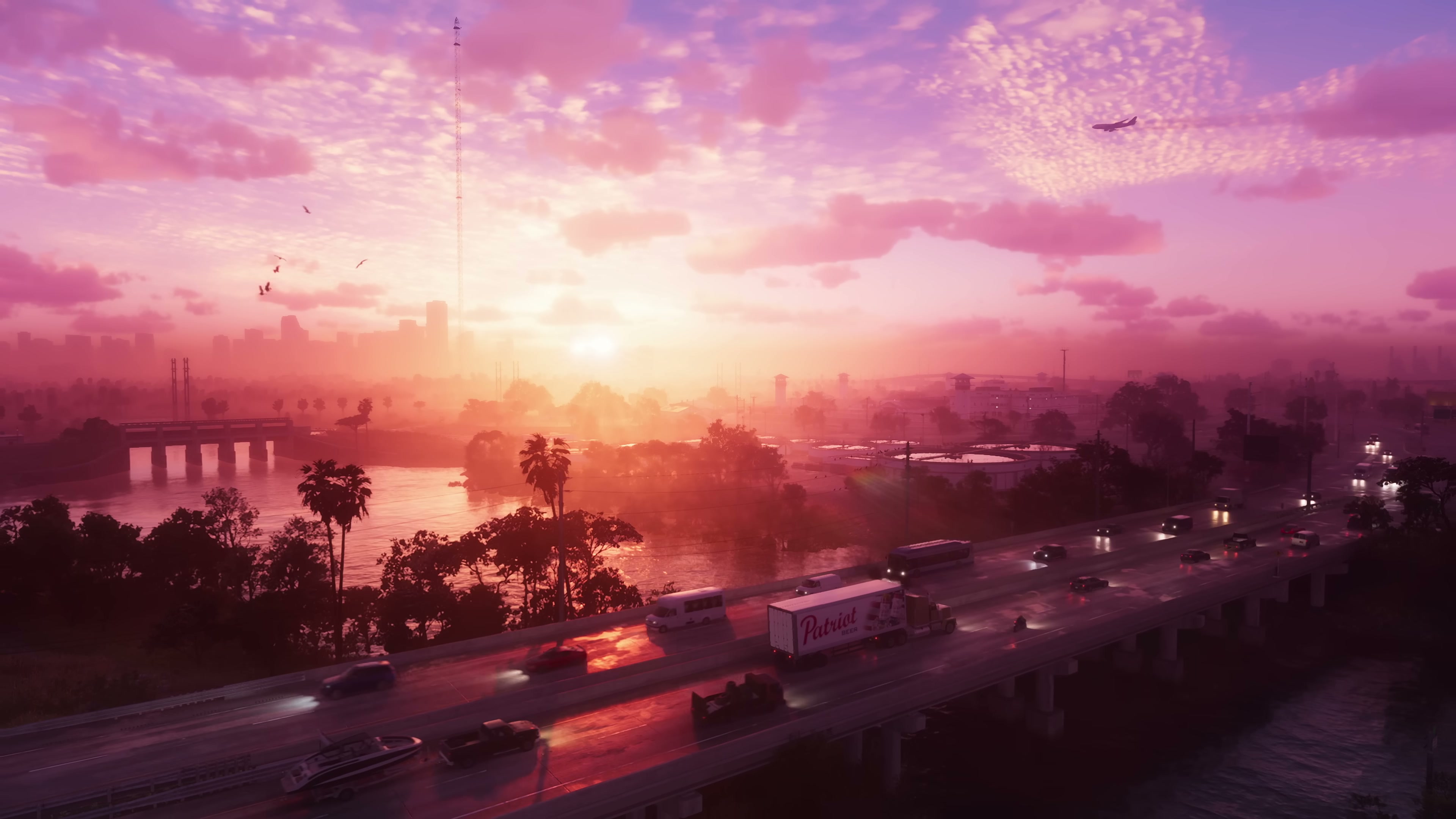 Aerial view of a multi-lane highway crossing water at sunset, with a Patriot semi-truck, palm trees, distant city skyline, and a pink-orange sky.