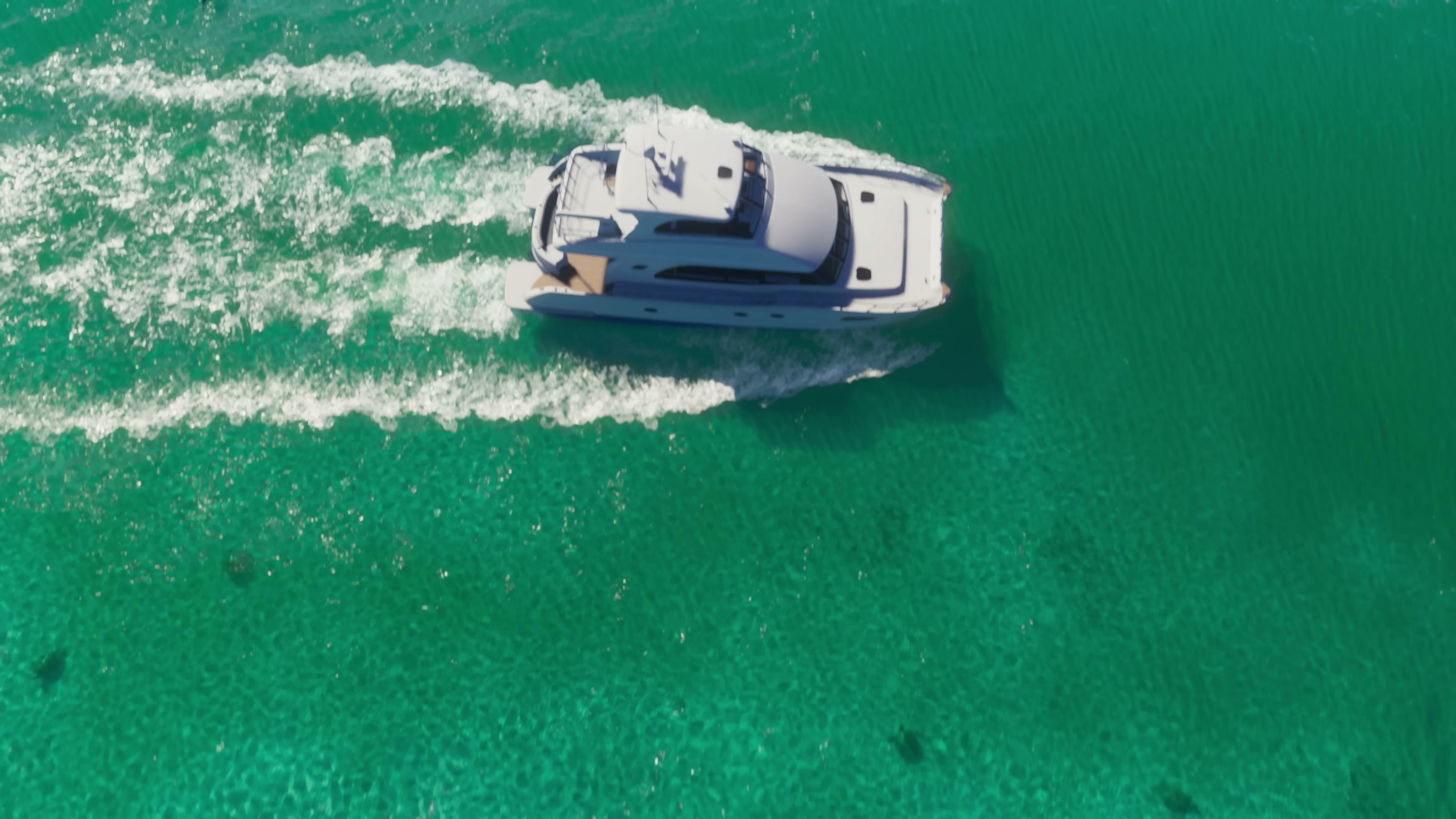 Aerial shot of a white yacht cutting through turquoise shallow water with visible sandy seabed and twin wake trails.