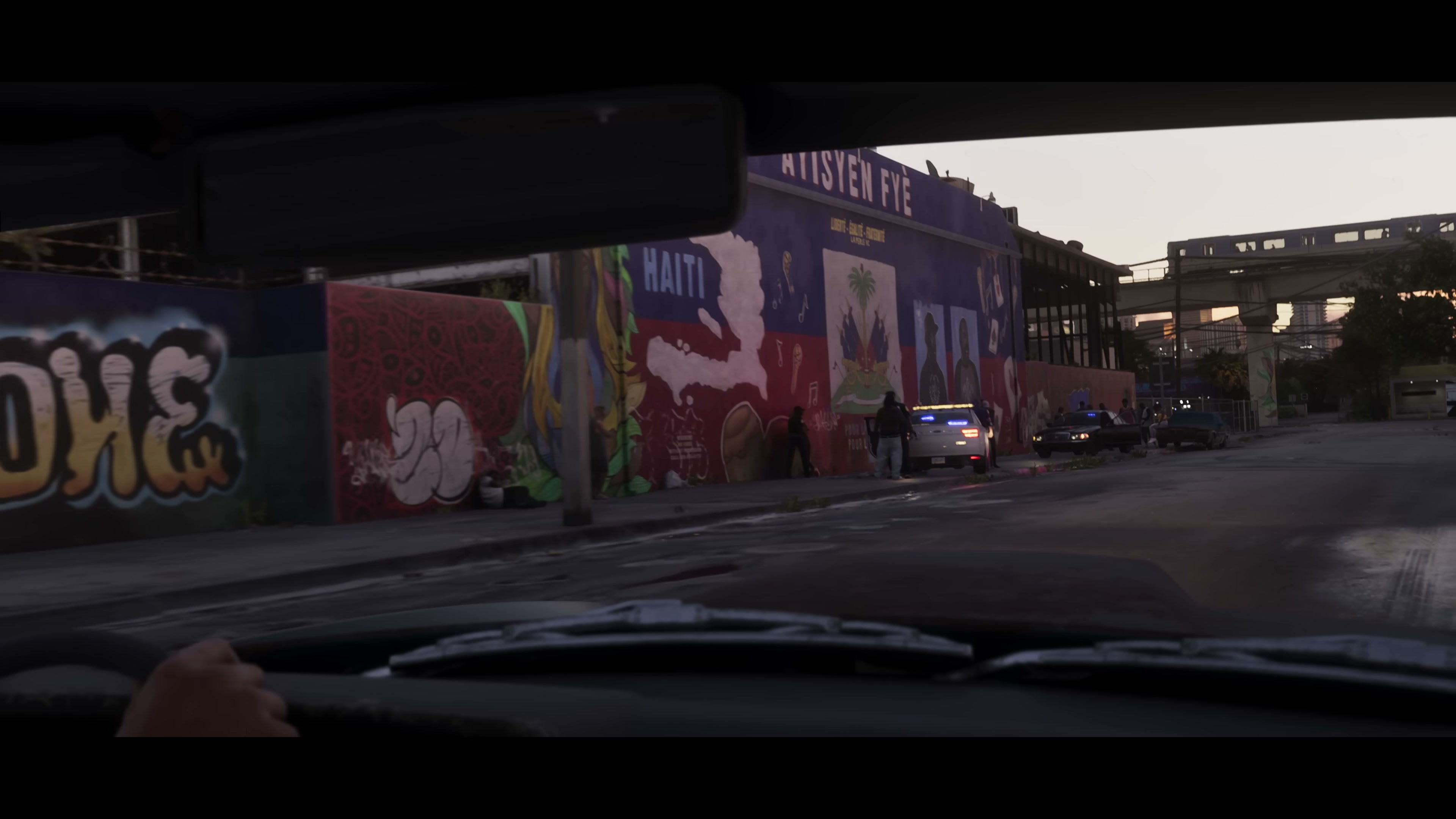 First-person view from a car interior with a driver's hand on the wheel passing an officer-led arrest beside a Haitian mural