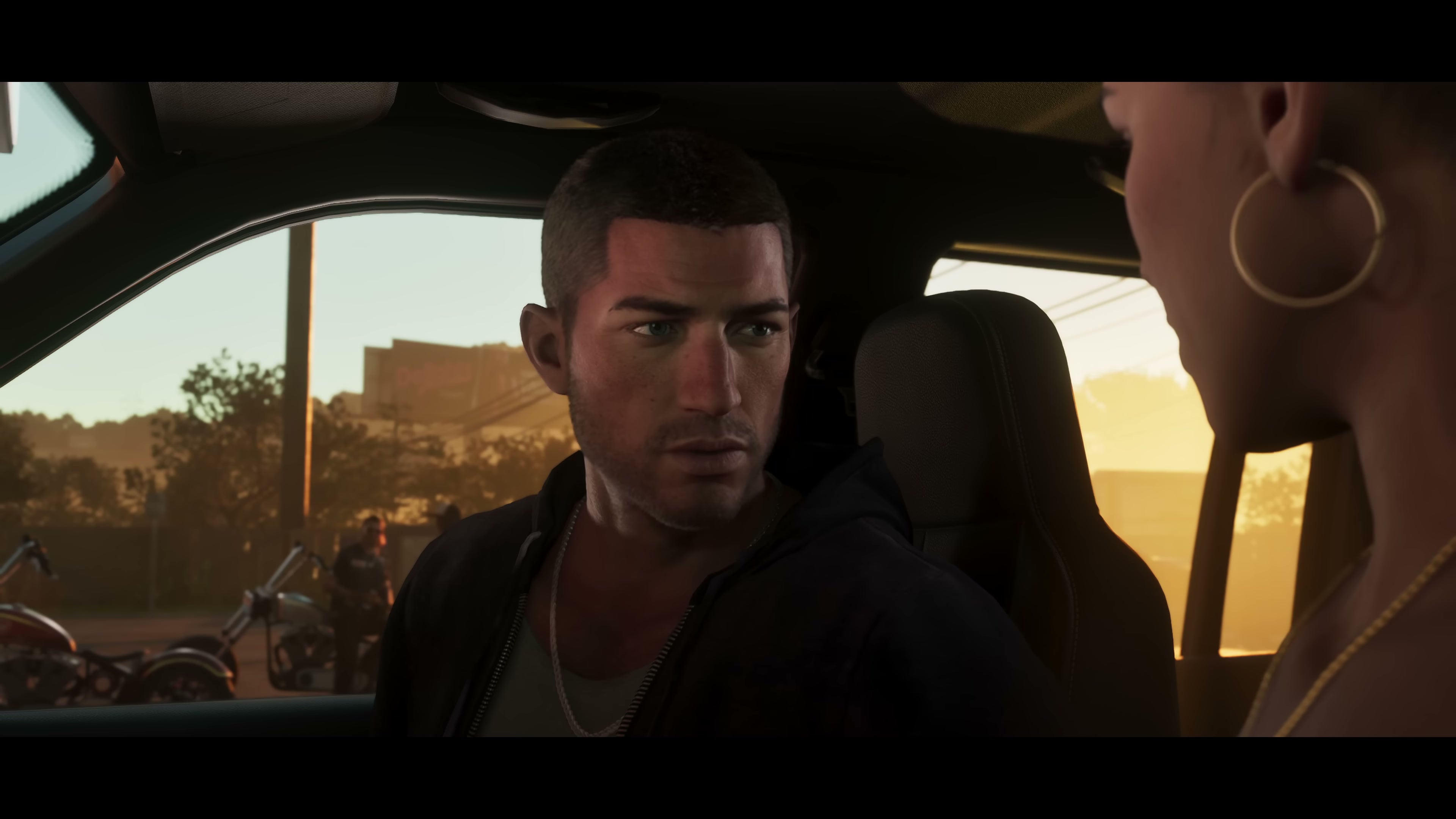 Jason in a dark hoodie and chain necklace looks toward Lucia from inside a vehicle at sunset.